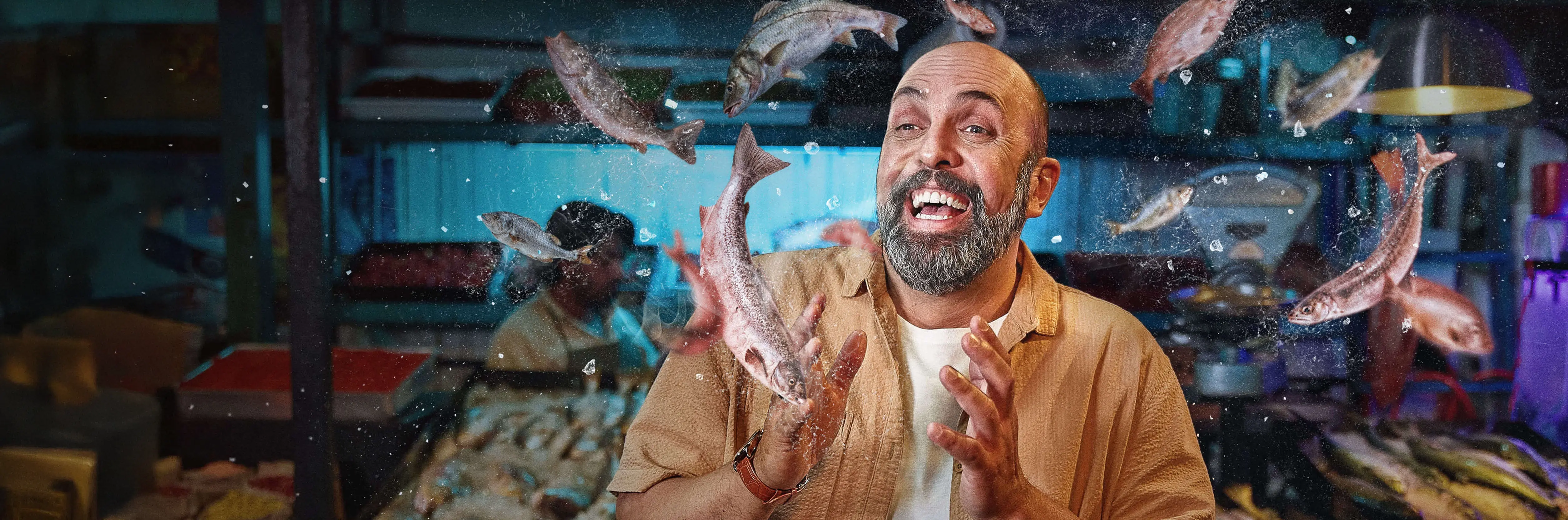 A laughing bearded man at a fish market, surrounded by an assortment of fish in the air.