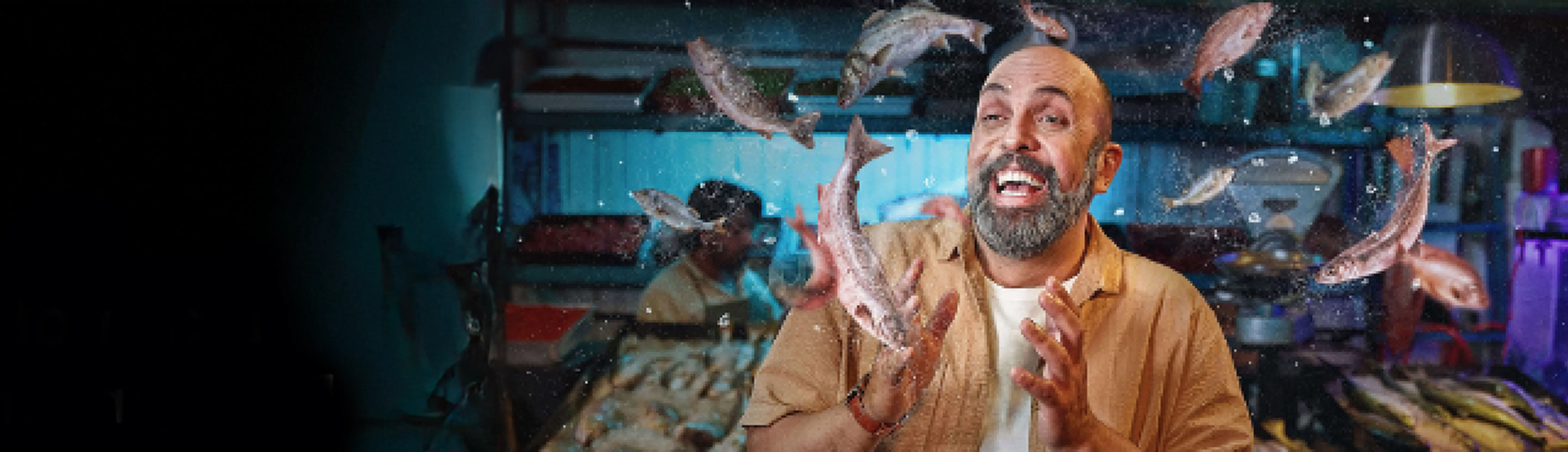 Un hombre barbudo y risueño en un mercado de pescado, rodeado de un surtido de peces en el aire.