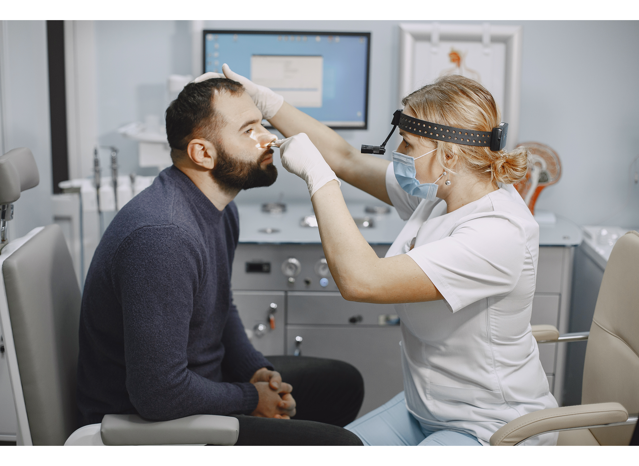 A healthcare provider wearing a face mask, gloves, and a head mounted light examines a male patient's nose in a medical exam room.