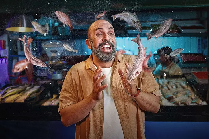 Un hombre barbudo y risueño en un mercado de pescado, rodeado de un surtido de peces en el aire.